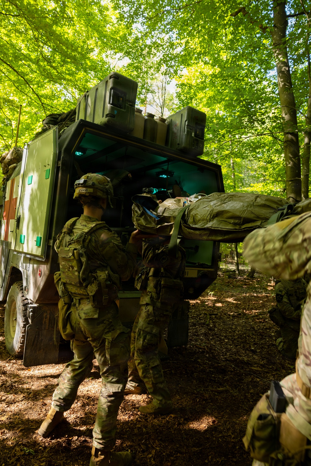 Military personnel in camouflage uniforms are loading a casualty on a litter/stretcher into the rear of a military medical vehicle (ambulance), identifiable by the red cross markings on its side. The scene takes place in a forested area with dense green tree canopy, suggesting a field exercise or operational environment. At least two soldiers are actively engaged in the litter-loading process.