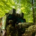 Military personnel in camouflage uniforms are loading a casualty on a litter/stretcher into the rear of a military medical vehicle (ambulance), identifiable by the red cross markings on its side. The scene takes place in a forested area with dense green tree canopy, suggesting a field exercise or operational environment. At least two soldiers are actively engaged in the litter-loading process.