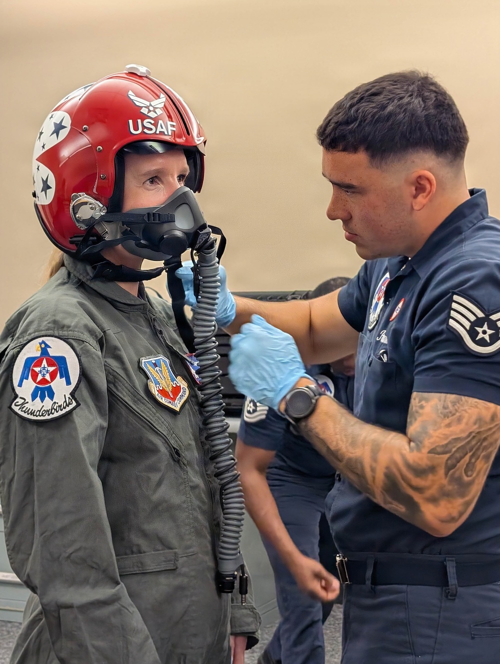 A USAF Thunderbirds crew member wearing blue latex gloves assists a person in a flight suit with fitting or adjusting an oxygen mask connected to a corrugated hose. The person being assisted wears a red USAF helmet with stars and a Thunderbirds patch on their flight suit, along with an Air Combat Command patch. The technician, wearing a dark blue Thunderbirds uniform with Staff Sergeant chevrons and visible arm tattoos, is focused on securing the mask connections.