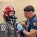 A USAF Thunderbirds crew member wearing blue latex gloves assists a person in a flight suit with fitting or adjusting an oxygen mask connected to a corrugated hose. The person being assisted wears a red USAF helmet with stars and a Thunderbirds patch on their flight suit, along with an Air Combat Command patch. The technician, wearing a dark blue Thunderbirds uniform with Staff Sergeant chevrons and visible arm tattoos, is focused on securing the mask connections.