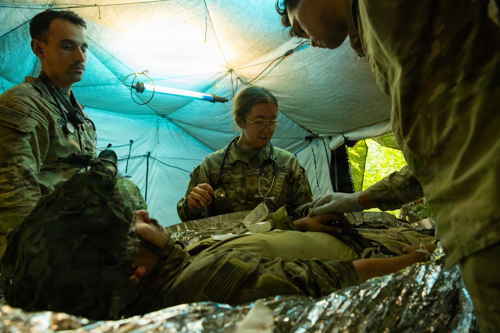 Military medical personnel are conducting a field medical treatment exercise inside a tactical tent shelter. Three soldiers in camouflage uniforms are attending to a simulated casualty lying on a surface covered with a mylar emergency blanket. The scene appears to be a training exercise based on the controlled environment and deliberate positioning of personnel.
