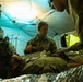 Military medical personnel are conducting a field medical treatment exercise inside a tactical tent shelter. Three soldiers in camouflage uniforms are attending to a simulated casualty lying on a surface covered with a mylar emergency blanket. The scene appears to be a training exercise based on the controlled environment and deliberate positioning of personnel.