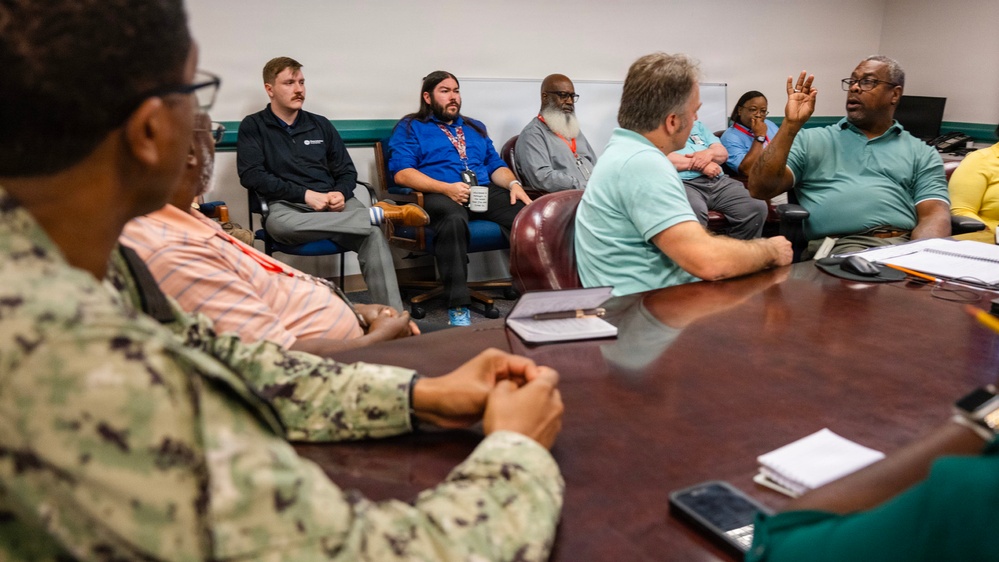 A formal meeting or briefing is taking place in a conference room with approximately 8-10 participants seated around and near a large wooden table. At least one individual is wearing U.S. Navy digital camouflage (NWU) uniform, while others are in civilian business casual attire. One participant on the right side of the table has a raised hand, suggesting active discussion or a question is being posed.