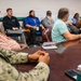 A formal meeting or briefing is taking place in a conference room with approximately 8-10 participants seated around and near a large wooden table. At least one individual is wearing U.S. Navy digital camouflage (NWU) uniform, while others are in civilian business casual attire. One participant on the right side of the table has a raised hand, suggesting active discussion or a question is being posed.