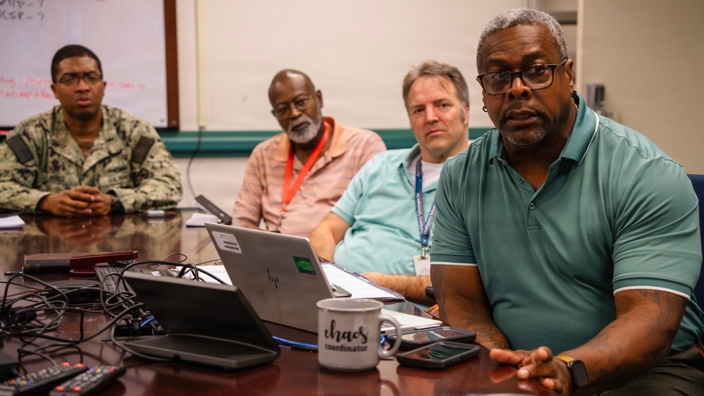 Four men are seated at a conference table in what appears to be a military or government meeting room. One individual on the left is wearing a U.S. Navy camouflage uniform, while the others are in civilian attire. The setting suggests an official briefing or coordination meeting.