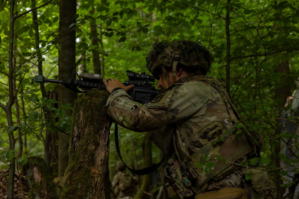 A soldier in full camouflage gear is positioned behind a moss-covered tree in a dense forest, aiming a rifle with an optical scope attachment. The soldier appears to be in a tactical training exercise or field operation, using the tree as cover while scanning downrange. A second soldier is faintly visible in the background.