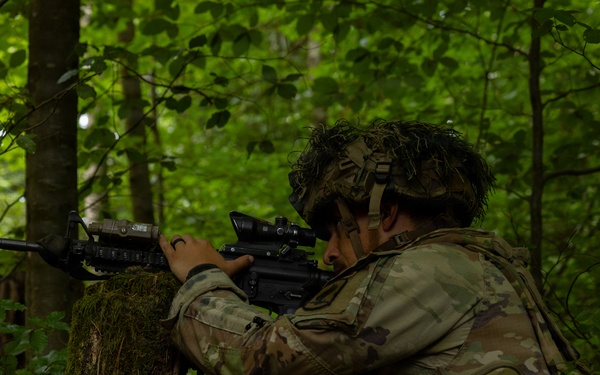 A soldier in full camouflage gear is positioned behind a moss-covered tree in a dense forest, aiming a rifle with an optical scope attachment. The soldier appears to be in a tactical training exercise or field operation, using the tree as cover while scanning downrange. A second soldier is faintly visible in the background.