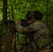 A soldier in full camouflage gear is positioned behind a moss-covered tree in a dense forest, aiming a rifle with an optical scope attachment. The soldier appears to be in a tactical training exercise or field operation, using the tree as cover while scanning downrange. A second soldier is faintly visible in the background.