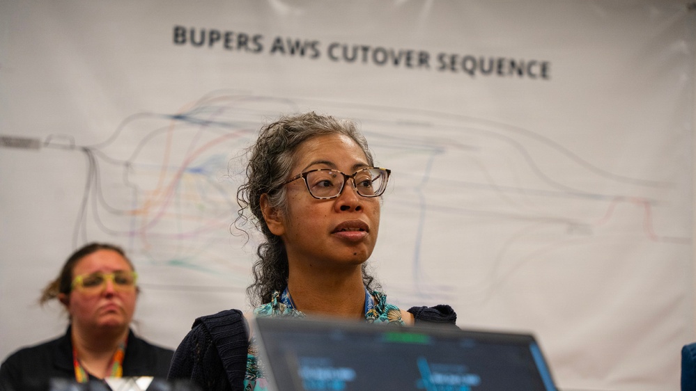 A woman wearing glasses and a floral top speaks or presents in front of a large banner reading 'BUPERS AWS CUTOVER SEQUENCE' with a technical diagram visible behind her. A second person is partially visible in the background to the left. The setting appears to be a professional or government IT operations briefing or command center environment.