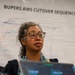 A woman wearing glasses and a floral top speaks or presents in front of a large banner reading 'BUPERS AWS CUTOVER SEQUENCE' with a technical diagram visible behind her. A second person is partially visible in the background to the left. The setting appears to be a professional or government IT operations briefing or command center environment.