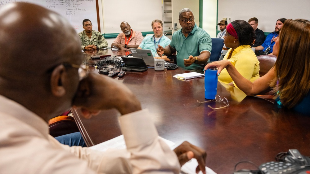 A group of approximately 10-12 people are seated around a large wooden conference table in what appears to be a formal meeting or briefing room. A man in a teal/green polo shirt appears to be the primary speaker, gesturing with his hands while addressing the group. The setting suggests a professional or government/military meeting environment, indicated by the presence of at least one person in military uniform.