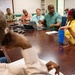 A group of approximately 10-12 people are seated around a large wooden conference table in what appears to be a formal meeting or briefing room. A man in a teal/green polo shirt appears to be the primary speaker, gesturing with his hands while addressing the group. The setting suggests a professional or government/military meeting environment, indicated by the presence of at least one person in military uniform.