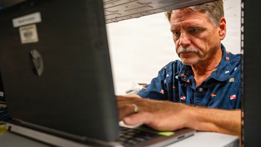 A middle-aged man with gray hair and a mustache is focused on working at an HP laptop computer. The photo is taken from a low angle through a shelf or rack opening, creating a framed perspective. He is wearing a navy blue shirt with small flag patterns.