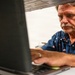 A middle-aged man with gray hair and a mustache is focused on working at an HP laptop computer. The photo is taken from a low angle through a shelf or rack opening, creating a framed perspective. He is wearing a navy blue shirt with small flag patterns.