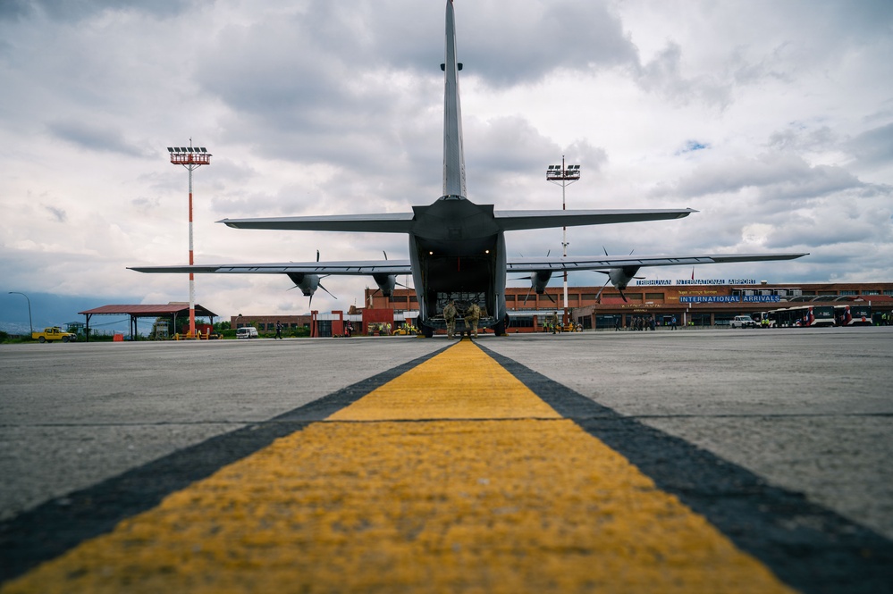 A military transport aircraft, likely a C-130 Hercules, is photographed from ground level at the rear, with its cargo ramp open and at least one uniformed personnel visible beneath it. The image is taken from a low angle along a yellow taxiway centerline at what is identified as Tribhuvan International Airport, visible in the background. The dramatic wide-angle perspective emphasizes the scale of the aircraft against an overcast sky.