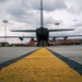 A military transport aircraft, likely a C-130 Hercules, is photographed from ground level at the rear, with its cargo ramp open and at least one uniformed personnel visible beneath it. The image is taken from a low angle along a yellow taxiway centerline at what is identified as Tribhuvan International Airport, visible in the background. The dramatic wide-angle perspective emphasizes the scale of the aircraft against an overcast sky.