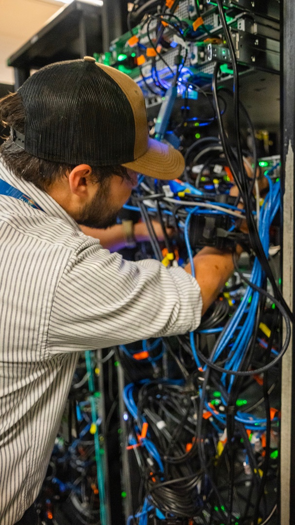 A male technician wearing a striped shirt and two-tone baseball cap is working inside a densely populated server rack or network cabinet. He is manually handling cables within a complex infrastructure setup featuring numerous blue, black, and other colored cables along with active networking equipment displaying green and blue indicator lights. The environment appears to be a data center or server room.