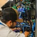 A male technician wearing a striped shirt and two-tone baseball cap is working inside a densely populated server rack or network cabinet. He is manually handling cables within a complex infrastructure setup featuring numerous blue, black, and other colored cables along with active networking equipment displaying green and blue indicator lights. The environment appears to be a data center or server room.
