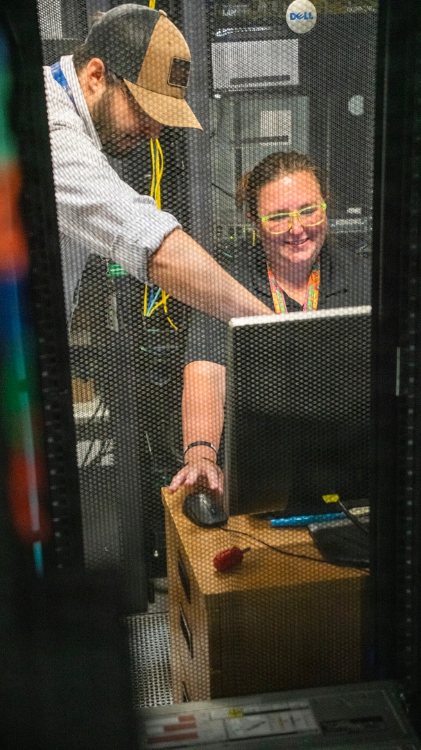 Two IT technicians are working inside or near a server rack enclosure, visible through a perforated mesh door. One person is operating a computer mouse while the other looks on at a monitor, suggesting collaborative troubleshooting or configuration work. The environment is clearly a data center with Dell-branded server equipment visible in the background.