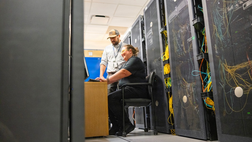 Two IT professionals are working together in a data center environment surrounded by server racks and cable management systems. One person is seated at a small workstation using a laptop while the other stands nearby, both appearing engaged and smiling. The scene depicts a collaborative technical work session amid dense networking infrastructure.