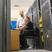 Two IT professionals are working together in a data center environment surrounded by server racks and cable management systems. One person is seated at a small workstation using a laptop while the other stands nearby, both appearing engaged and smiling. The scene depicts a collaborative technical work session amid dense networking infrastructure.