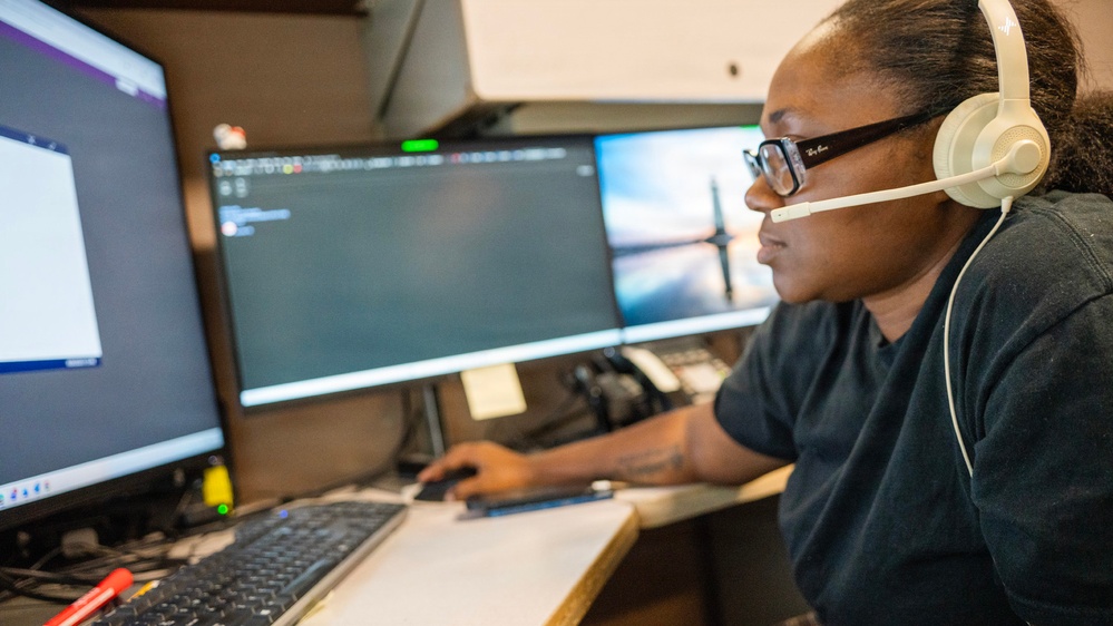 A person wearing white headphones and Ray-Ban glasses is seated at a multi-monitor workstation, actively working at a computer. The setup includes at least three monitors displaying various screens, including what appears to be a dark-themed interface or terminal. The environment suggests a professional or technical workspace such as a dispatch center, call center, or IT operations room.