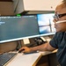 A person wearing white headphones and Ray-Ban glasses is seated at a multi-monitor workstation, actively working at a computer. The setup includes at least three monitors displaying various screens, including what appears to be a dark-themed interface or terminal. The environment suggests a professional or technical workspace such as a dispatch center, call center, or IT operations room.