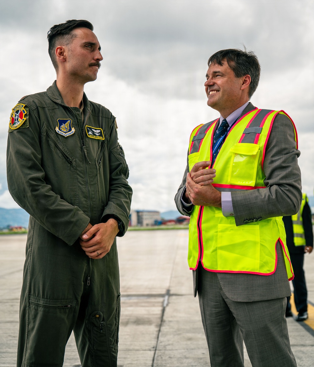 Two men are standing on an airport tarmac or airfield ramp engaged in conversation. One is wearing a military flight suit with patches indicating Pacific Air Forces affiliation, while the other is dressed in a business suit with a high-visibility safety vest. The civilian appears to be smiling and looking upward, suggesting a positive interaction.