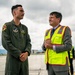 Two men are standing on an airport tarmac or airfield ramp engaged in conversation. One is wearing a military flight suit with patches indicating Pacific Air Forces affiliation, while the other is dressed in a business suit with a high-visibility safety vest. The civilian appears to be smiling and looking upward, suggesting a positive interaction.
