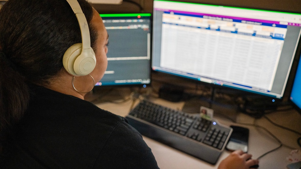 A person with dark hair is seated at a workstation with multiple monitors, wearing cream/beige over-ear headphones and a hoop earring. They appear to be working in a professional environment, possibly a call center, dispatch center, or data operations role. The dual-monitor setup displays what appears to be scheduling, dispatch, or database software with colorful interface elements.