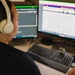 A person with dark hair is seated at a workstation with multiple monitors, wearing cream/beige over-ear headphones and a hoop earring. They appear to be working in a professional environment, possibly a call center, dispatch center, or data operations role. The dual-monitor setup displays what appears to be scheduling, dispatch, or database software with colorful interface elements.