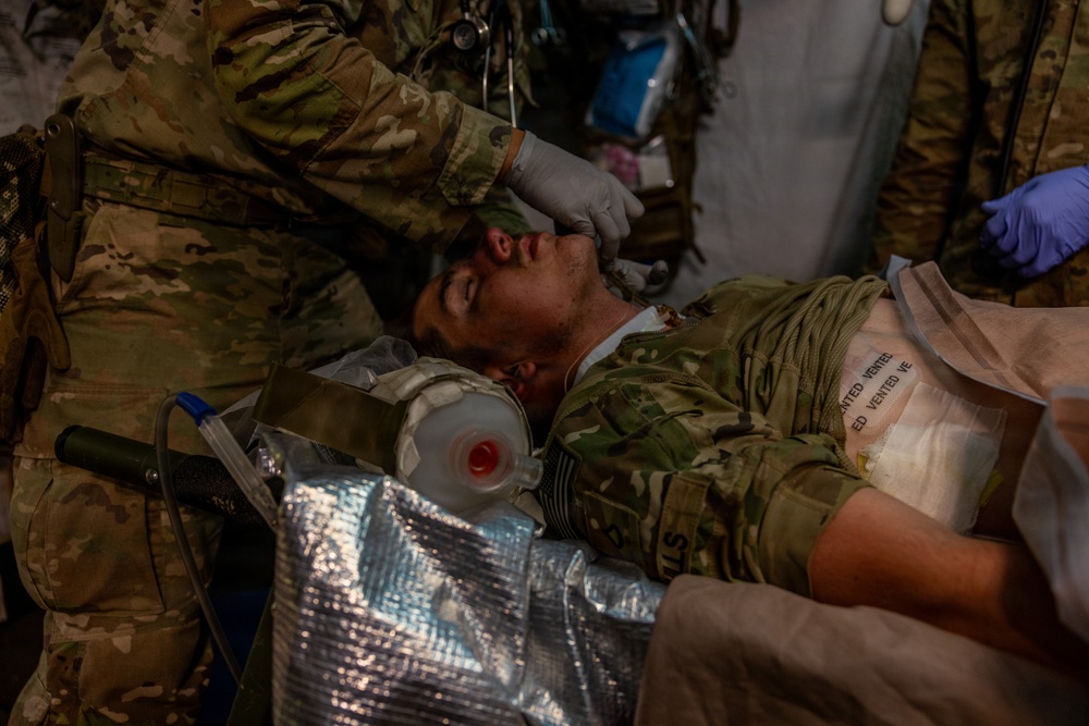 Military medical personnel in camouflage uniforms are providing emergency care to a casualty lying on a stretcher. The patient is wearing military combat uniform and has medical equipment including a breathing mask and vented chest seals applied. Multiple providers wearing gloves are simultaneously attending to the patient, suggesting a trauma scenario or training exercise.