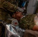 Military medical personnel in camouflage uniforms are providing emergency care to a casualty lying on a stretcher. The patient is wearing military combat uniform and has medical equipment including a breathing mask and vented chest seals applied. Multiple providers wearing gloves are simultaneously attending to the patient, suggesting a trauma scenario or training exercise.