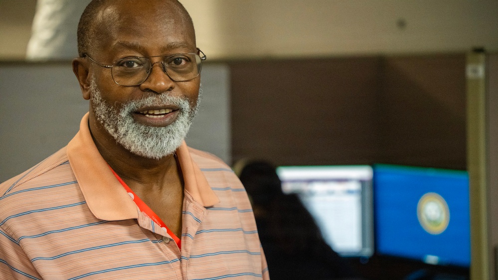 A middle-aged man with a gray beard and glasses is smiling toward the camera in what appears to be an office or workplace environment. He is wearing a peach/salmon striped polo shirt and a red lanyard. In the background, computer monitors are visible, one displaying what appears to be a state seal on a blue background.
