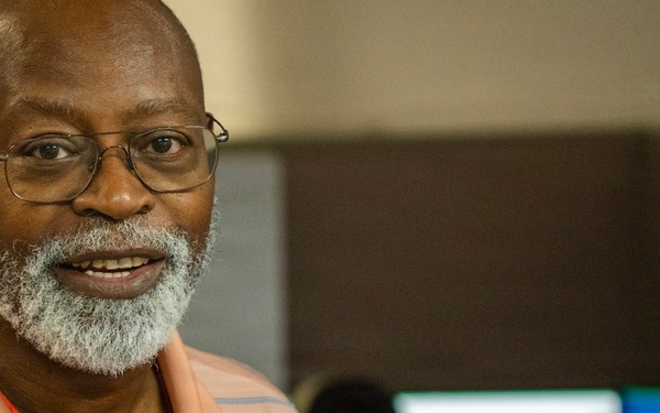 A middle-aged man with a gray beard and glasses is smiling toward the camera in what appears to be an office or workplace environment. He is wearing a peach/salmon striped polo shirt and a red lanyard. In the background, computer monitors are visible, one displaying what appears to be a state seal on a blue background.