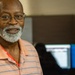 A middle-aged man with a gray beard and glasses is smiling toward the camera in what appears to be an office or workplace environment. He is wearing a peach/salmon striped polo shirt and a red lanyard. In the background, computer monitors are visible, one displaying what appears to be a state seal on a blue background.