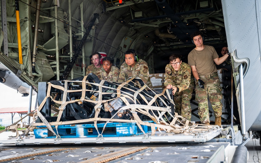 Military personnel in camouflage uniforms are loading or unloading cargo from the rear ramp of a military transport aircraft, likely a C-130 Hercules. A netted cargo pallet on a blue military pallet base is being pushed or positioned on the aircraft's roller-equipped cargo floor. The group consists of approximately five to six service members, some in full uniform with gear and one in a plain tan t-shirt.