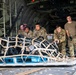Military personnel in camouflage uniforms are loading or unloading cargo from the rear ramp of a military transport aircraft, likely a C-130 Hercules. A netted cargo pallet on a blue military pallet base is being pushed or positioned on the aircraft's roller-equipped cargo floor. The group consists of approximately five to six service members, some in full uniform with gear and one in a plain tan t-shirt.