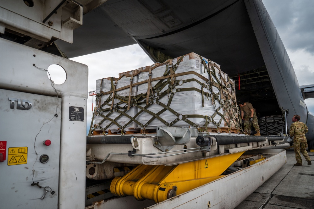 Military personnel are loading or unloading a large cargo pallet secured with olive-drab cargo netting onto or off a military transport aircraft via a ground loading platform. The pallet contains stacked white boxes or packages strapped tightly together. At least two uniformed service members in camouflage uniforms are visible overseeing or guiding the operation.