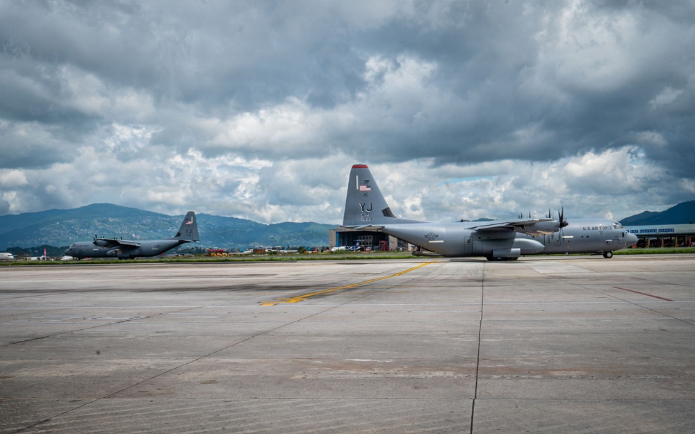 Two U.S. Air Force C-130J Super Hercules military transport aircraft are parked on a concrete flight line apron, likely at a forward operating base or joint-use airfield. The foreground aircraft displays tail code 'YJ' and 'U.S. AIR FORCE' markings, consistent with Yokota Air Base, Japan. Mountains and overcast skies are visible in the background.