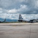 Two U.S. Air Force C-130J Super Hercules military transport aircraft are parked on a concrete flight line apron, likely at a forward operating base or joint-use airfield. The foreground aircraft displays tail code 'YJ' and 'U.S. AIR FORCE' markings, consistent with Yokota Air Base, Japan. Mountains and overcast skies are visible in the background.