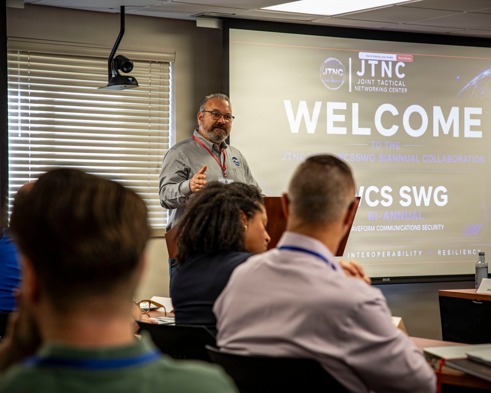 A presenter stands at the front of a conference room delivering a welcome address at the JTNC (Joint Tactical Networking Center) JTNC/WNCSSWG Biannual Collaboration event. The projected slide prominently displays 'WELCOME' along with references to WCS SWG (Waveform Communications Security Sub-Working Group), emphasizing themes of interoperability and resiliency. Several attendees are seated facing the presenter, with lanyards and name badges visible.