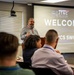 A presenter stands at the front of a conference room delivering a welcome address at the JTNC (Joint Tactical Networking Center) JTNC/WNCSSWG Biannual Collaboration event. The projected slide prominently displays 'WELCOME' along with references to WCS SWG (Waveform Communications Security Sub-Working Group), emphasizing themes of interoperability and resiliency. Several attendees are seated facing the presenter, with lanyards and name badges visible.