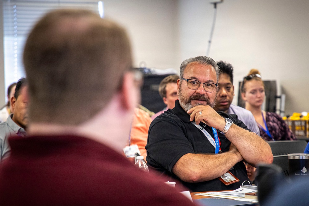 A group of adults are seated in what appears to be a conference room or training session. The central figure, a middle-aged man wearing glasses, a black polo shirt, and a blue lanyard, is attentively listening with his hand raised to his chin in a thoughtful pose. Several other attendees are visible in the background, slightly out of focus.