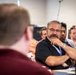 A group of adults are seated in what appears to be a conference room or training session. The central figure, a middle-aged man wearing glasses, a black polo shirt, and a blue lanyard, is attentively listening with his hand raised to his chin in a thoughtful pose. Several other attendees are visible in the background, slightly out of focus.