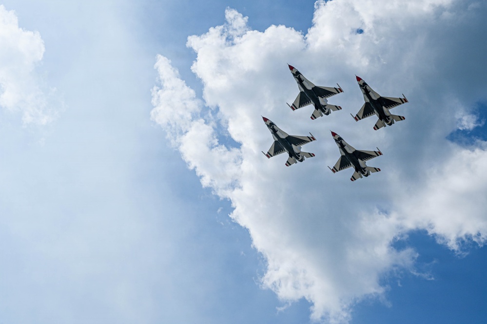 Four military jet aircraft are flying in a tight diamond formation against a partly cloudy blue sky, photographed from directly below. The aircraft display a distinctive white, red, and dark blue color scheme consistent with a precision aerobatic demonstration team. The low-angle upward perspective provides a dramatic view of the aircraft's undersides and wing configurations.