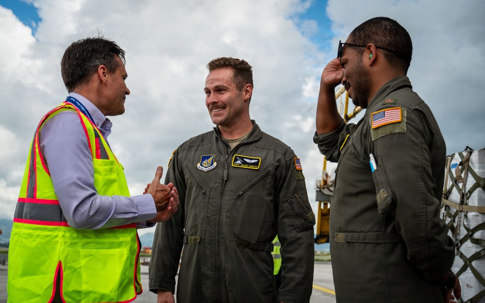 Two U.S. Air Force personnel in olive drab flight suits are greeting a civilian wearing a high-visibility safety vest on what appears to be an airport tarmac or flight line. The center airman, whose name tag reads 'CAPT JACOB CARLEY,' is shaking hands with the civilian while smiling. Cargo equipment and aircraft ground support machinery are visible in the background.