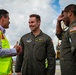 Two U.S. Air Force personnel in olive drab flight suits are greeting a civilian wearing a high-visibility safety vest on what appears to be an airport tarmac or flight line. The center airman, whose name tag reads 'CAPT JACOB CARLEY,' is shaking hands with the civilian while smiling. Cargo equipment and aircraft ground support machinery are visible in the background.