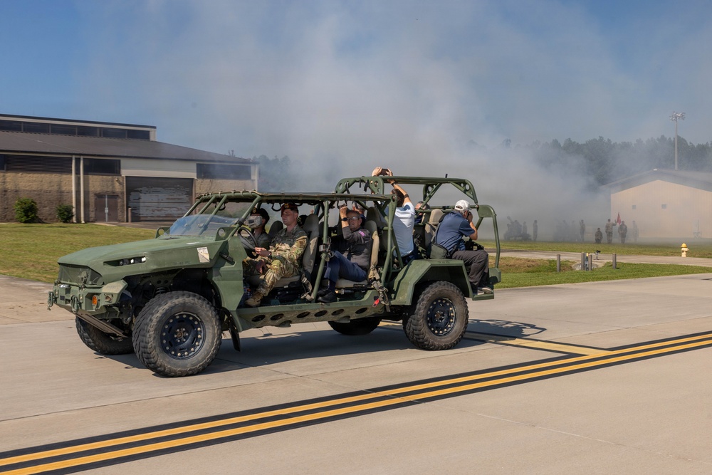 A military open-frame tactical vehicle, consistent with an Infantry Squad Vehicle (ISV), is driving on a paved road at what appears to be a military installation. The vehicle carries several occupants, including at least one person in military camouflage uniform and several civilians. Smoke or haze is visible in the background, suggesting a demonstration or exercise is underway.