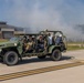 A military open-frame tactical vehicle, consistent with an Infantry Squad Vehicle (ISV), is driving on a paved road at what appears to be a military installation. The vehicle carries several occupants, including at least one person in military camouflage uniform and several civilians. Smoke or haze is visible in the background, suggesting a demonstration or exercise is underway.
